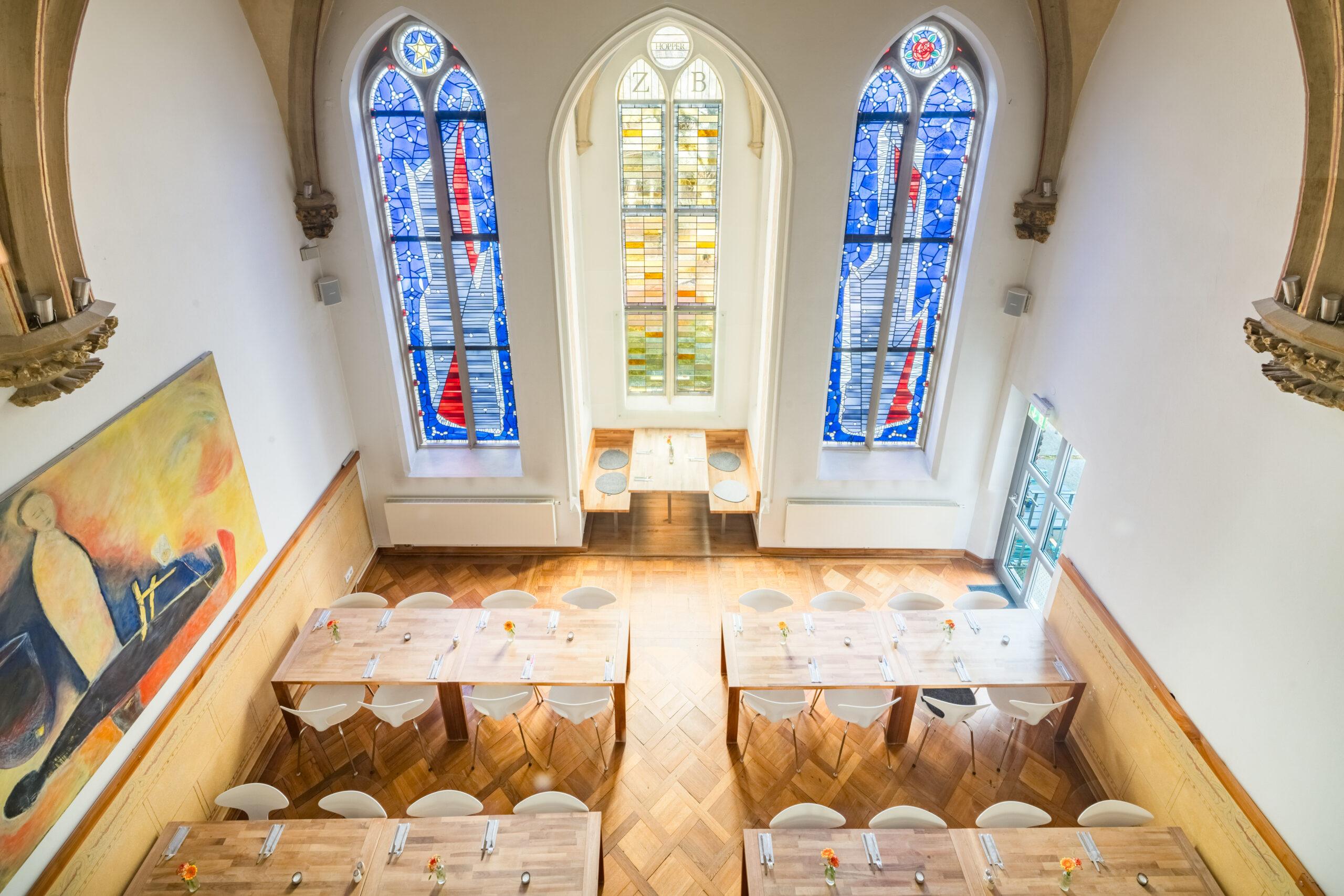 Heller Veranstaltungsraum mit Stuhlreihen und bunten Kirchenfenstern.